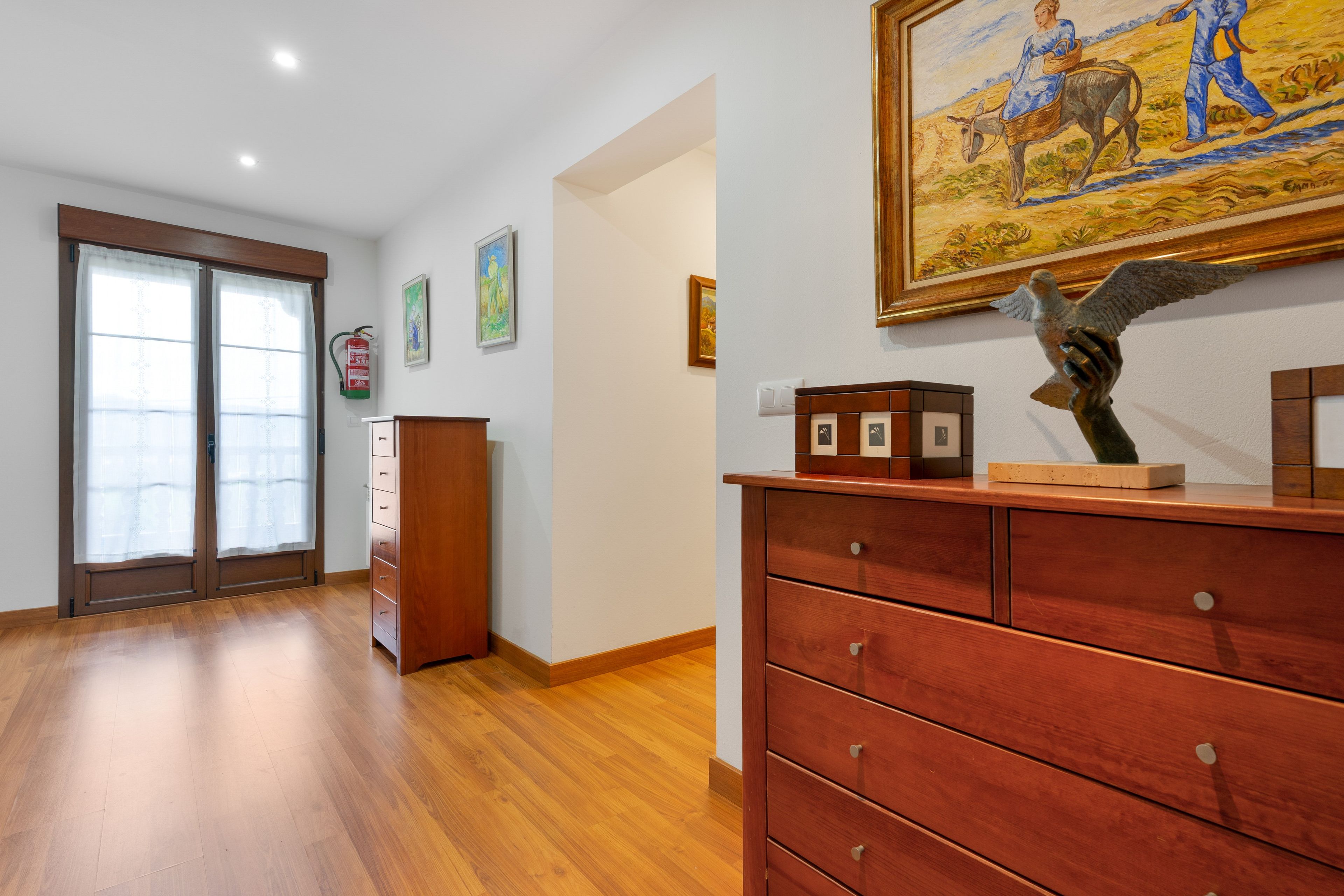 A hotel room with a wooden floor, a wooden dresser, a painting on the wall, and a fire extinguisher mounted on the wall.