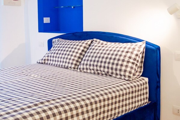A bed with a checkered bedspread and matching pillows, a blue headboard, and a wall-mounted shelf with a glass container. - [Cozy] Blue Velvet Loft* (Torino)