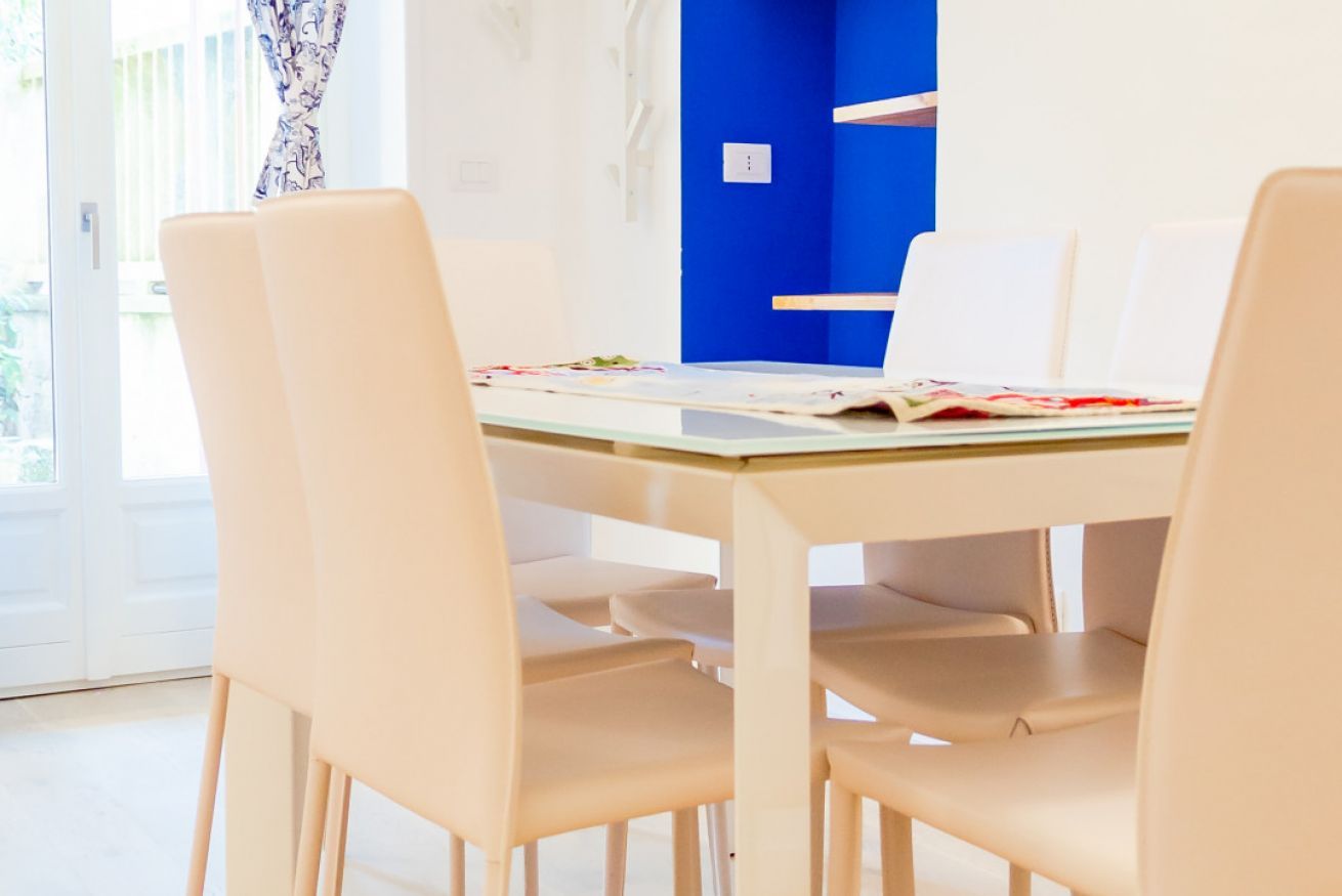 Un coin repas avec une table en verre et des chaises de couleur beige, un mur d’accent bleu et une vue à travers une porte vitrée.