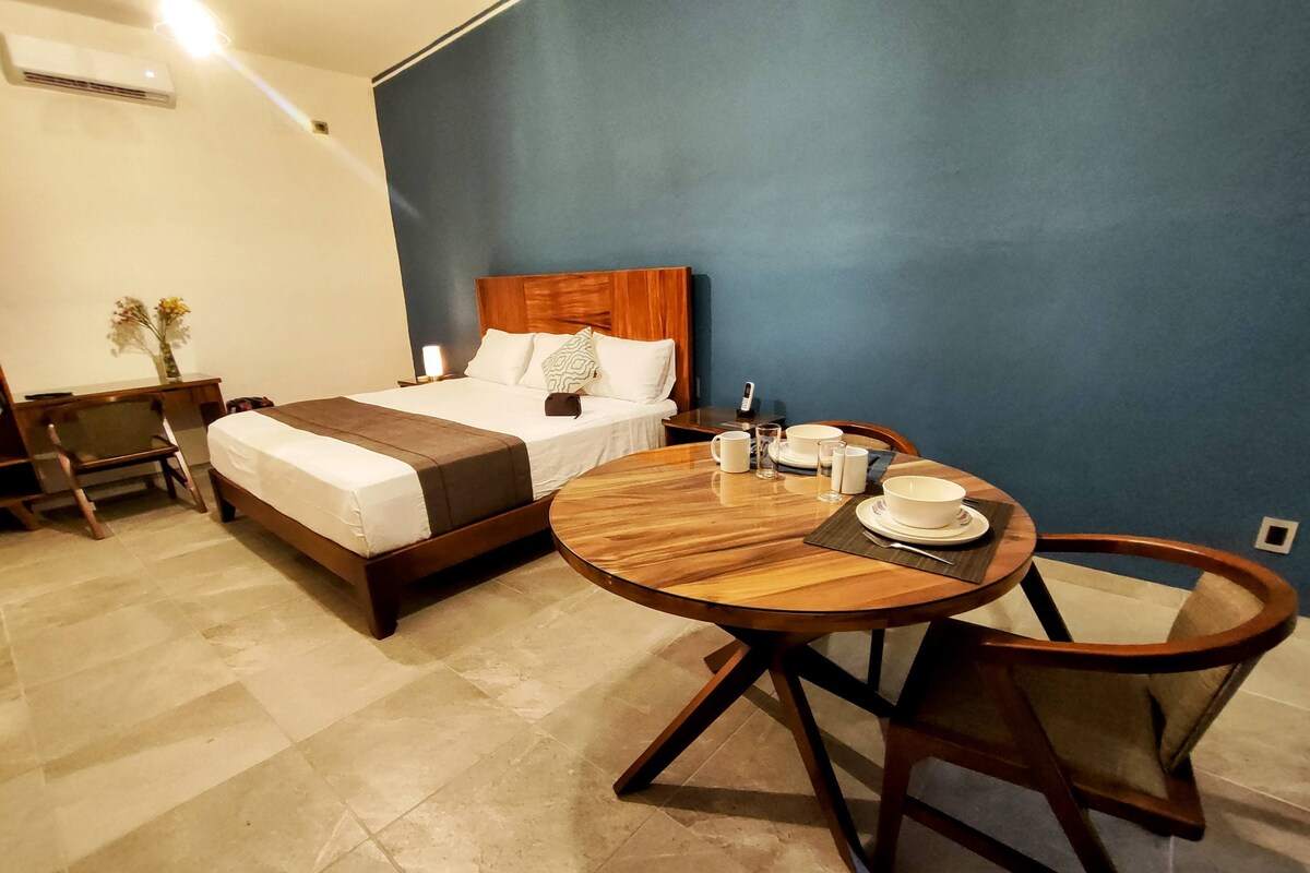 Quarto de hotel com cama, mesa redonda de madeira com cadeiras, mesa de cabeceira com abajur e um vaso com flores.