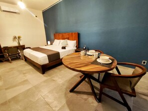 Quarto de hotel com cama, mesa redonda de madeira com cadeiras, mesa de cabeceira com abajur e um vaso com flores.