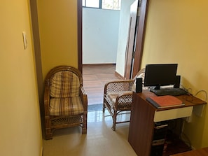 A room with a desk, two wicker chairs, a computer setup, and a door leading to another room.