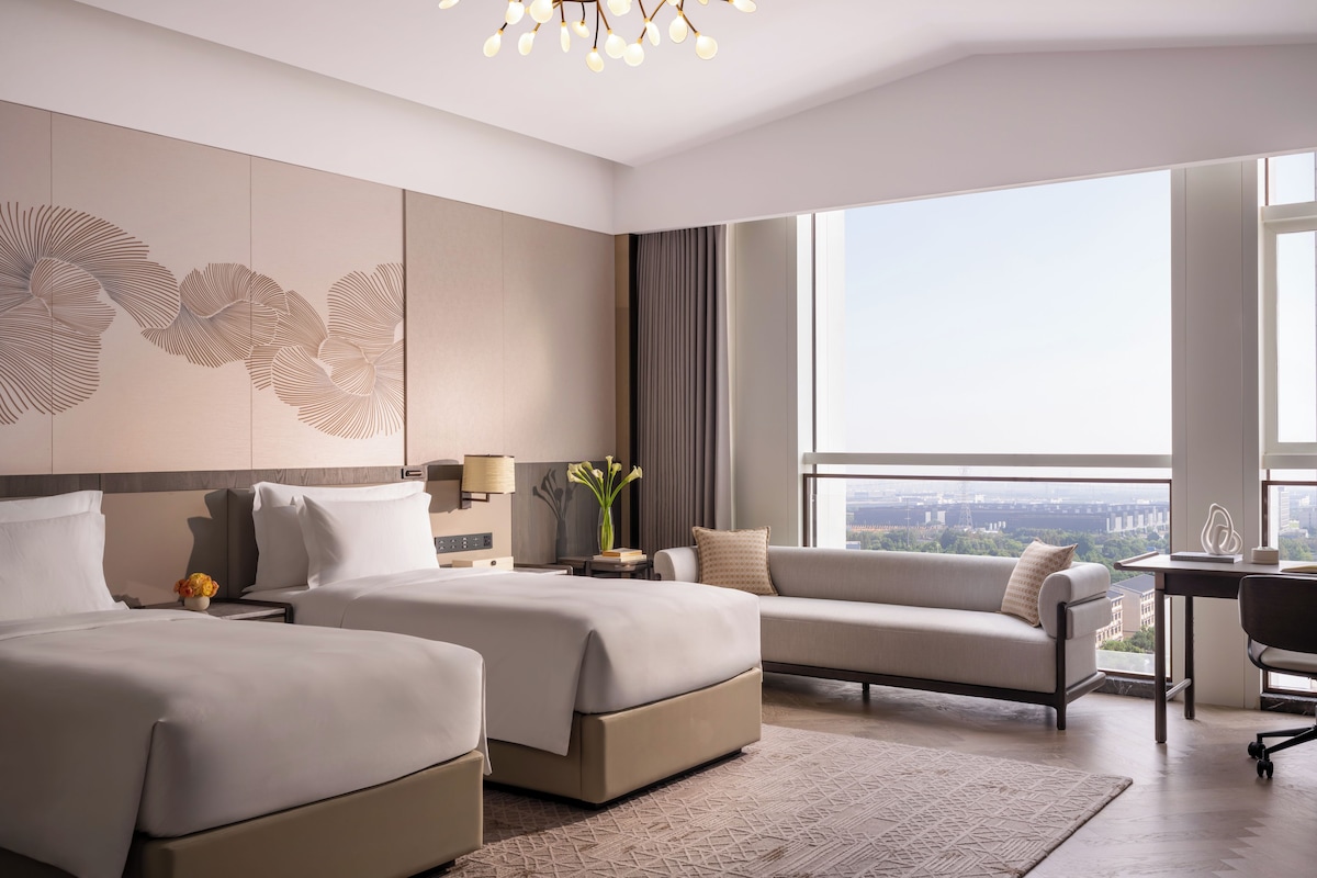 Habitación de hotel moderna con dos camas, un sofá, un escritorio y un amplio ventanal con vistas a la ciudad.