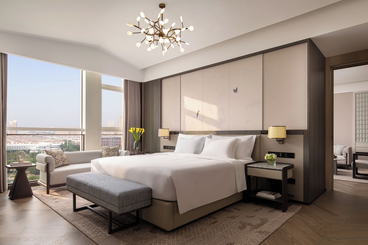 Una habitación de hotel moderna con una cama grande, una zona de estar, vistas de la ciudad y una lámpara decorativa.