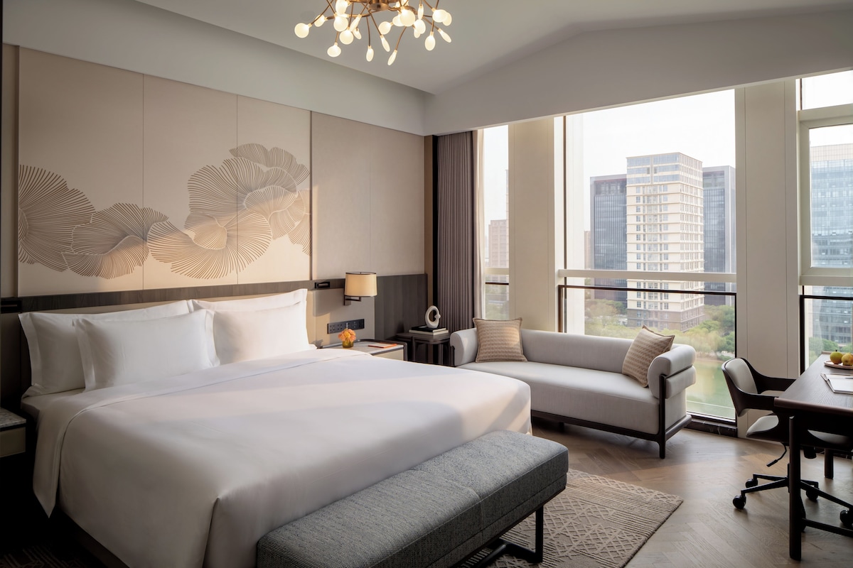 Habitación de hotel moderna con una cama grande, un sofá, un escritorio y vistas de la ciudad a través de amplias ventanas.