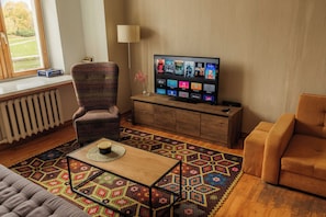 A living room with a television, a patterned rug, and a window with a view of trees. - Old Town Riverside Apartment, Retro Vibe & River Panorama (Vilnius)
