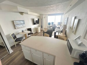 Een moderne keuken met een centraal eiland, een eethoek met stoelen, een flatscreen televisie aan de muur en een groot raam dat voor natuurlijk licht zorgt.
