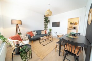 Ein modernes Wohnzimmer mit einer Couch, einem Couchtisch und einem Esstisch, einer hängenden Pflanze und einem Fernseher an der Wand.