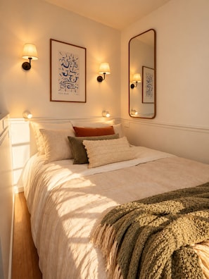 A neatly made bed with a beige comforter and a knitted throw blanket. There are two wall-mounted lamps with light shades and a framed abstract artwork above the bed. - New Listing (Paris)