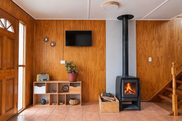 A room with a wooden interior, a television mounted on the wall, a wood-burning stove, and a staircase.