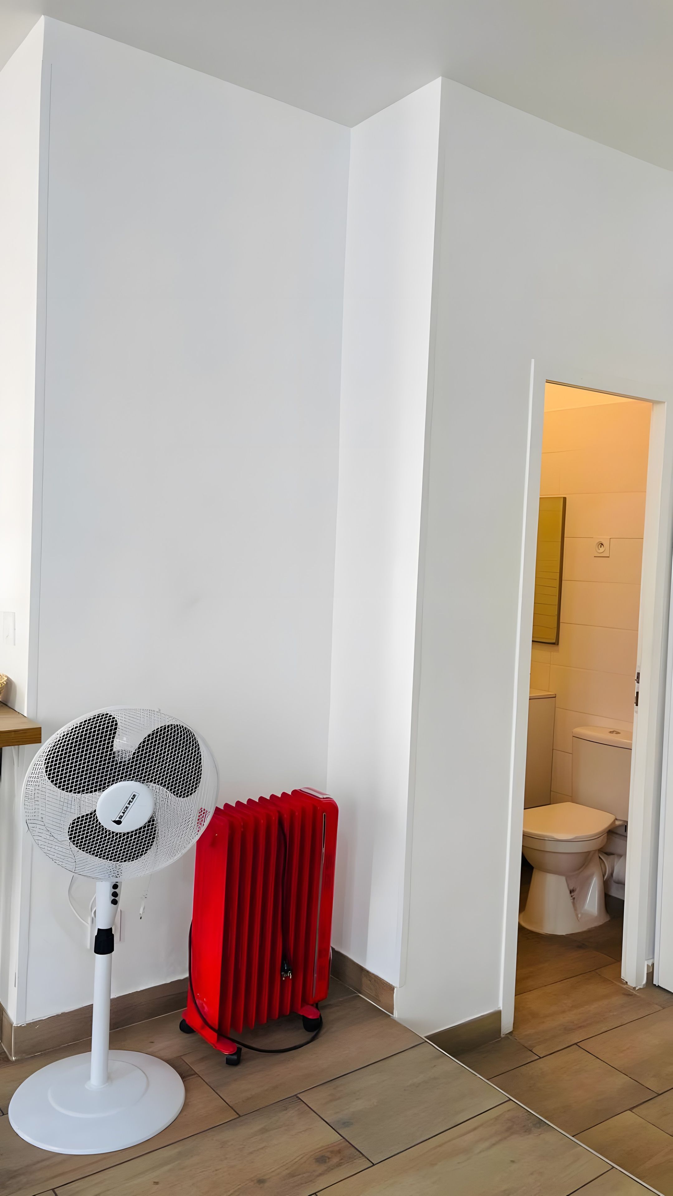 Une pièce avec un radiateur blanc, un ventilateur sur pied et une valise rouge.