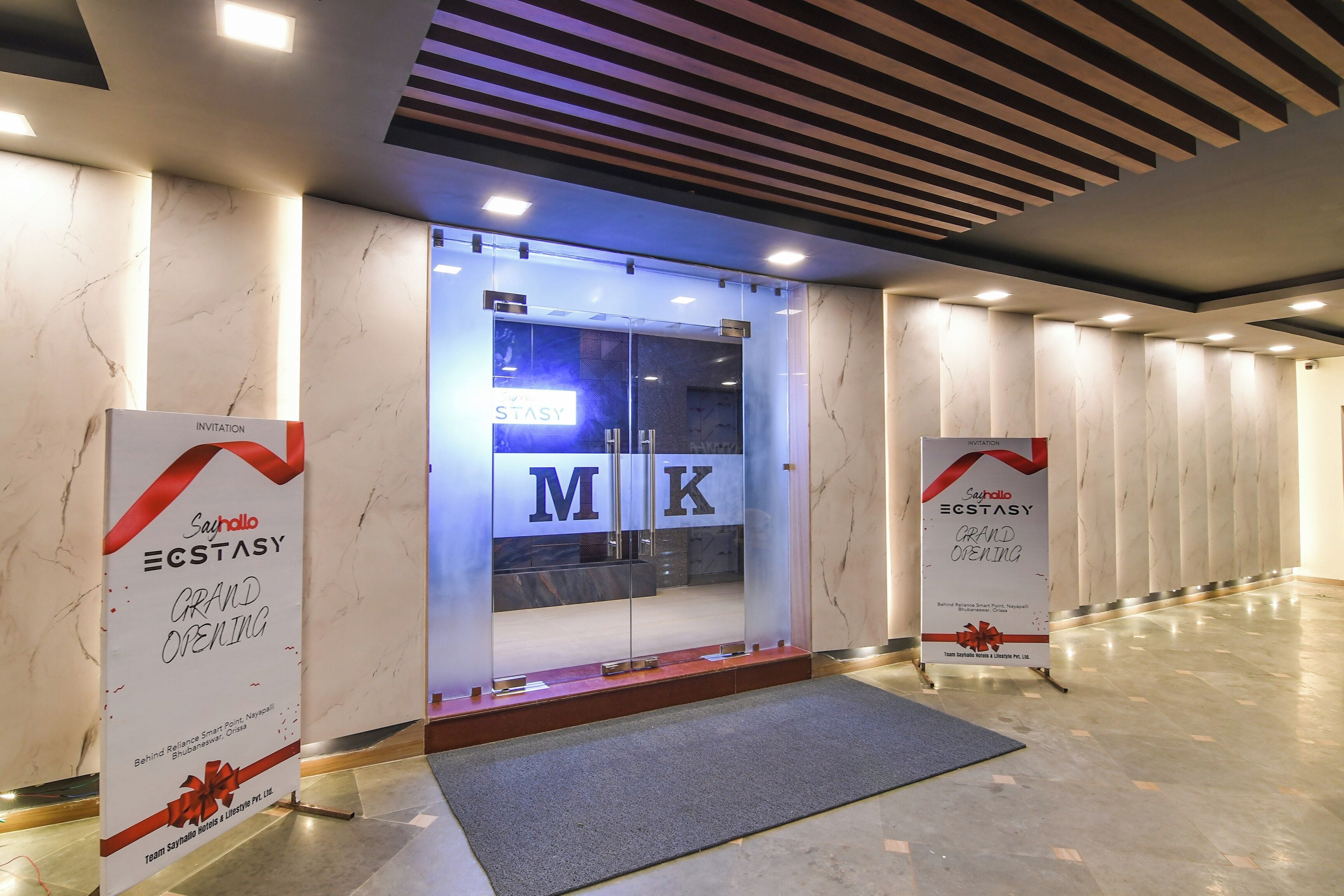 A hotel entrance with a modern design, featuring a glass door with the letters 'M K' and a sign indicating a grand opening.
