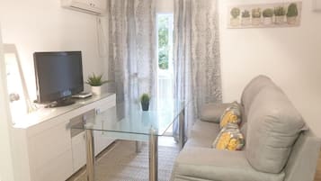Un espace de vie moderne comprenant une table à manger avec un plateau en verre, un canapé avec des coussins, un téléviseur à écran plat et une plante en pot.