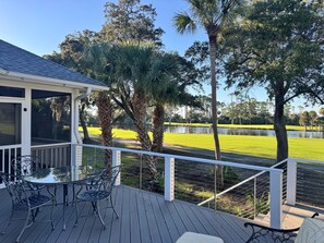 A covered outdoor deck with a glass table and chairs, overlooking a well-maintained lawn and a body of water. - Marsh View @ 162 (Fripp Island)