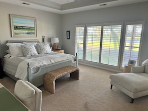 A bedroom with a bed, a bench, a chair, a nightstand, and a window with blinds. - Marsh View @ 162 (Fripp Island)