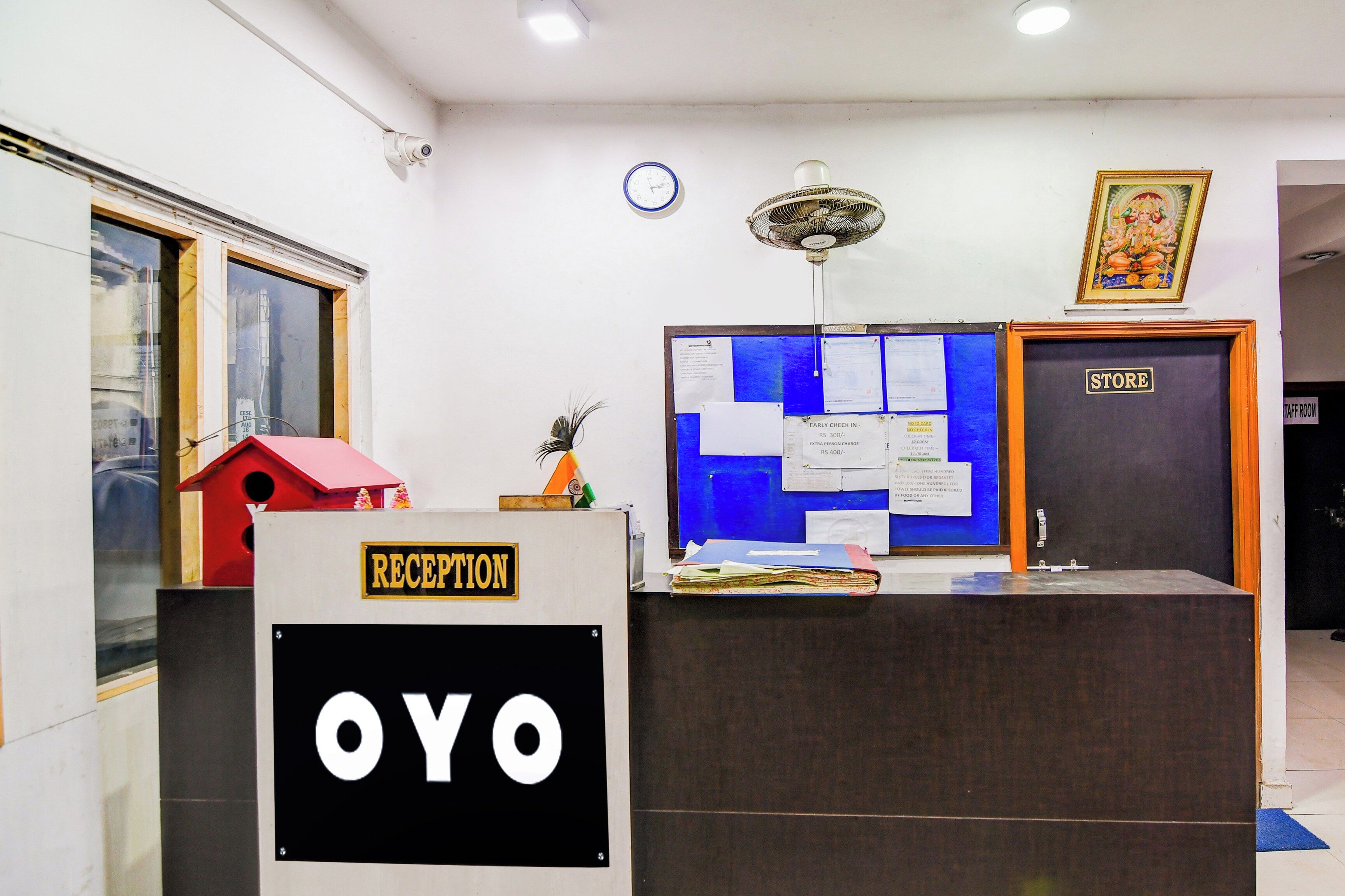 Una reception con un cartello che riporta 'RECEPTION' e 'OYO'. Si nota una bacheca blu con vari avvisi, un orologio e una ventola a soffitto sopra il bancone.