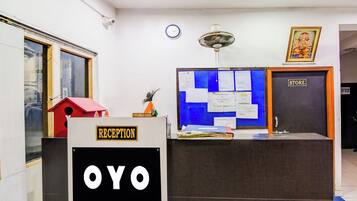 Un mostrador de recepción con un letrero que dice 'RECEPTION' y 'OYO'. Hay una pizarra azul con varios avisos, un reloj y un ventilador de techo sobre el mostrador.