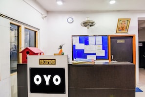 Un mostrador de recepción con un letrero que dice 'RECEPTION' y 'OYO'. Hay una pizarra azul con varios avisos, un reloj y un ventilador de techo sobre el mostrador.