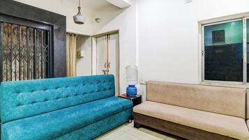 Una habitación con un sofá azul, un sofá color beige, un dispensador de agua, una lámpara en el techo y una puerta con reja de metal.