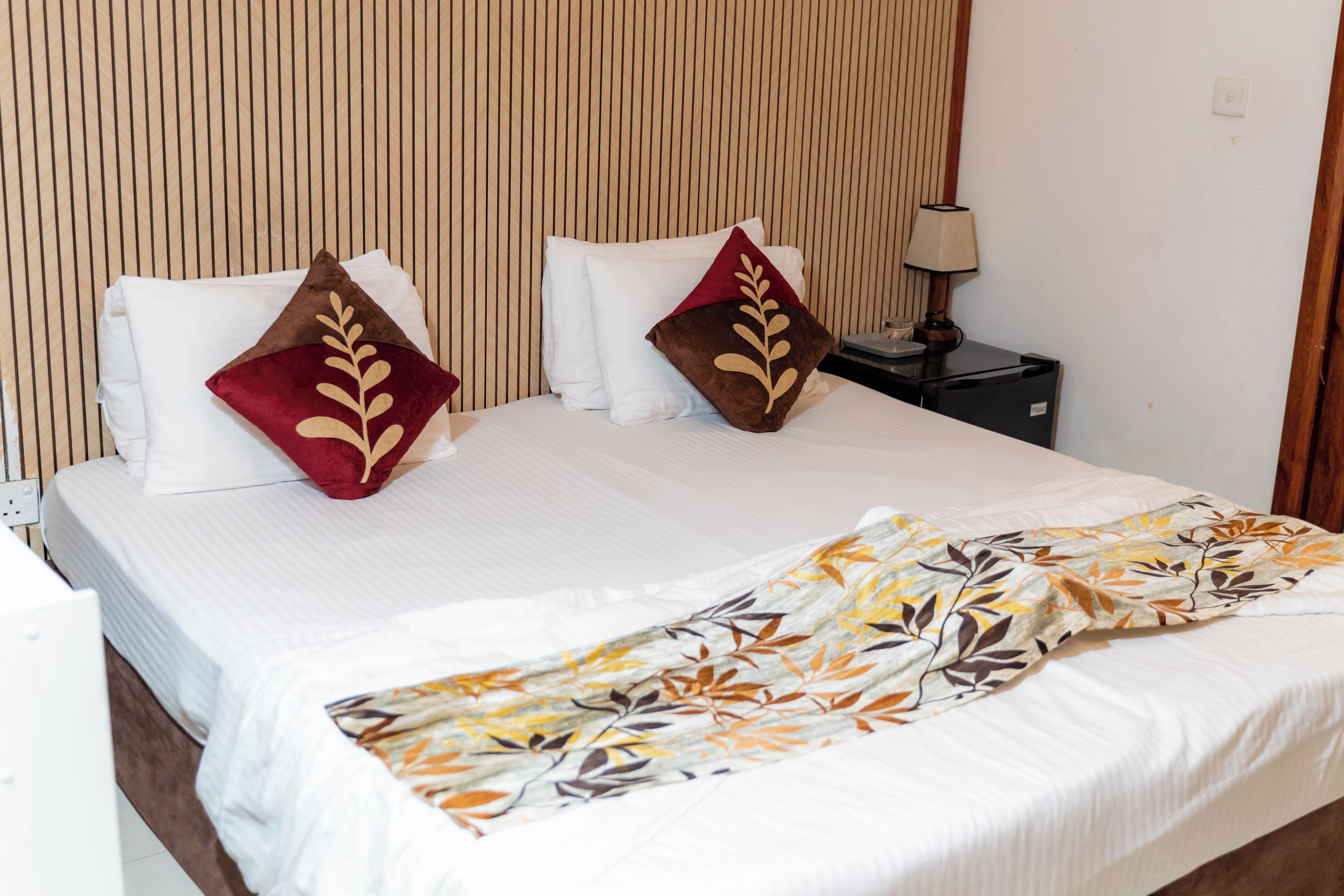 Una habitación de hotel con una cama, almohadas, una mesita de noche, una lámpara y un enchufe.