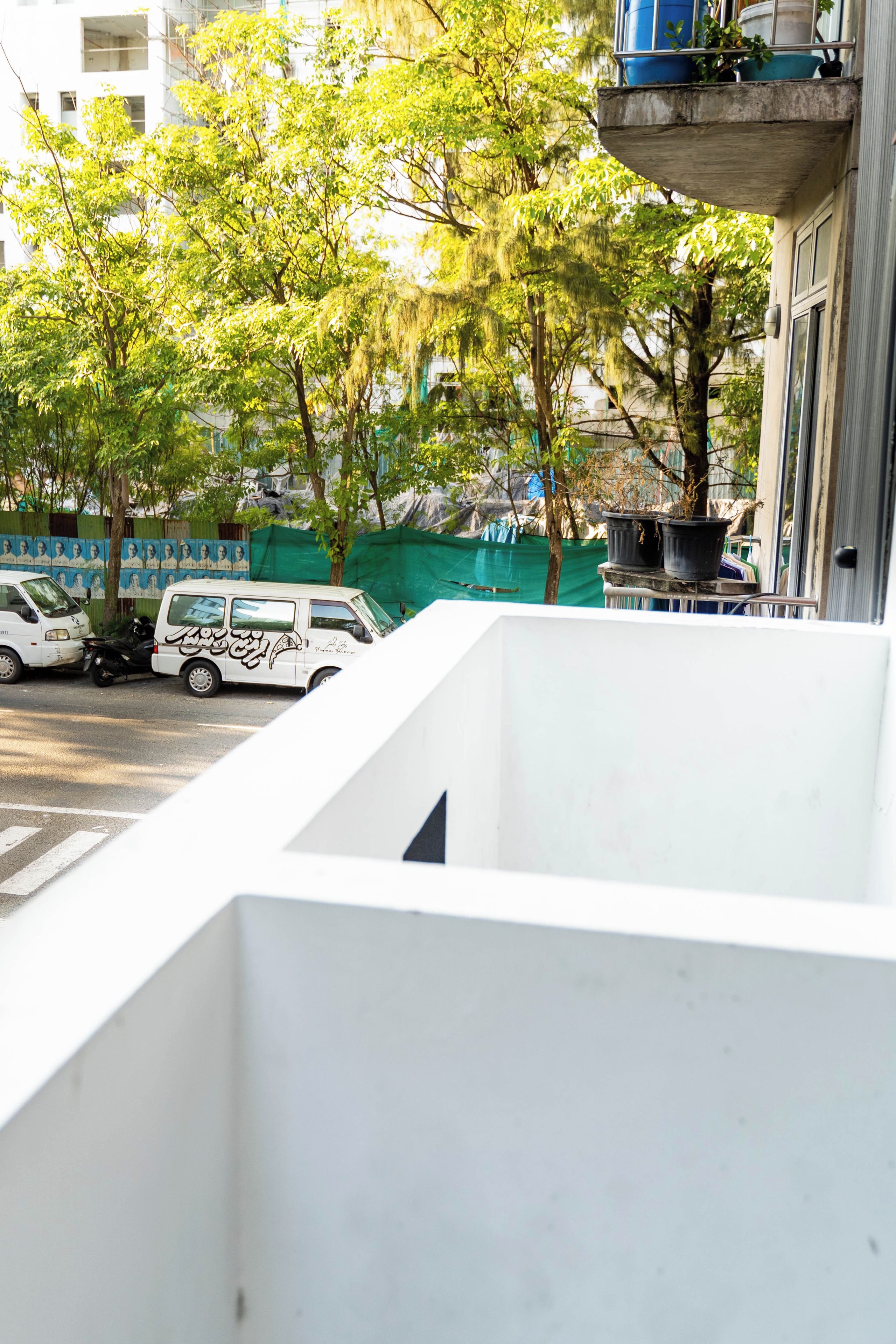 Un edificio blanco con balcón, una furgoneta blanca estacionada y una valla verde al fondo.