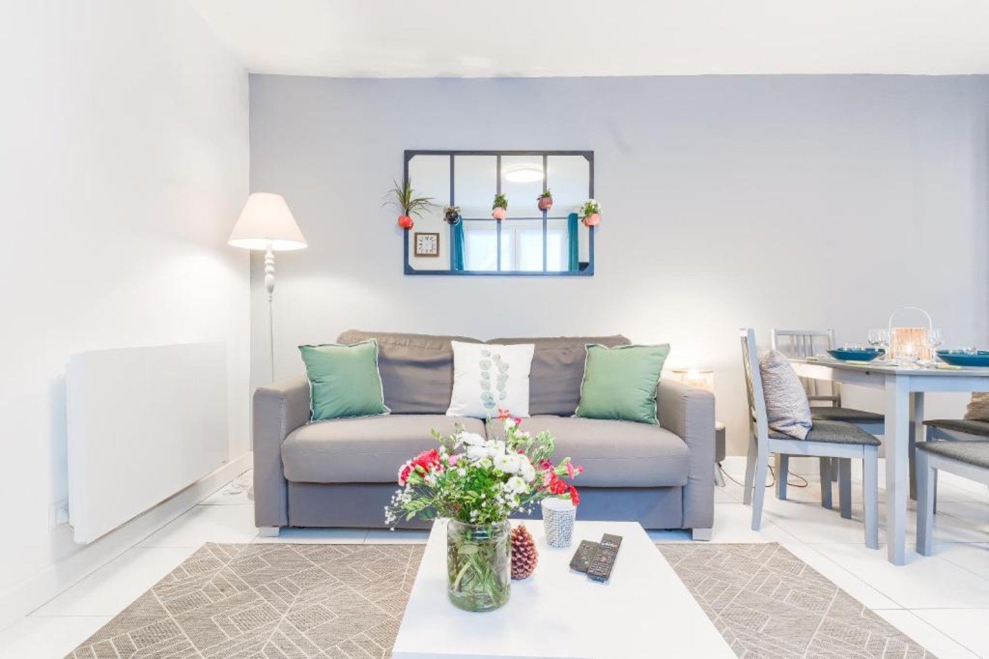 Een woonkamer met een grijze bank, een salontafel met een bloevas, een lamp en een eethoek met een tafel en stoelen.