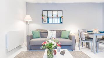 Een woonkamer met een grijze bank, een salontafel met een bloevas, een lamp en een eethoek met een tafel en stoelen.