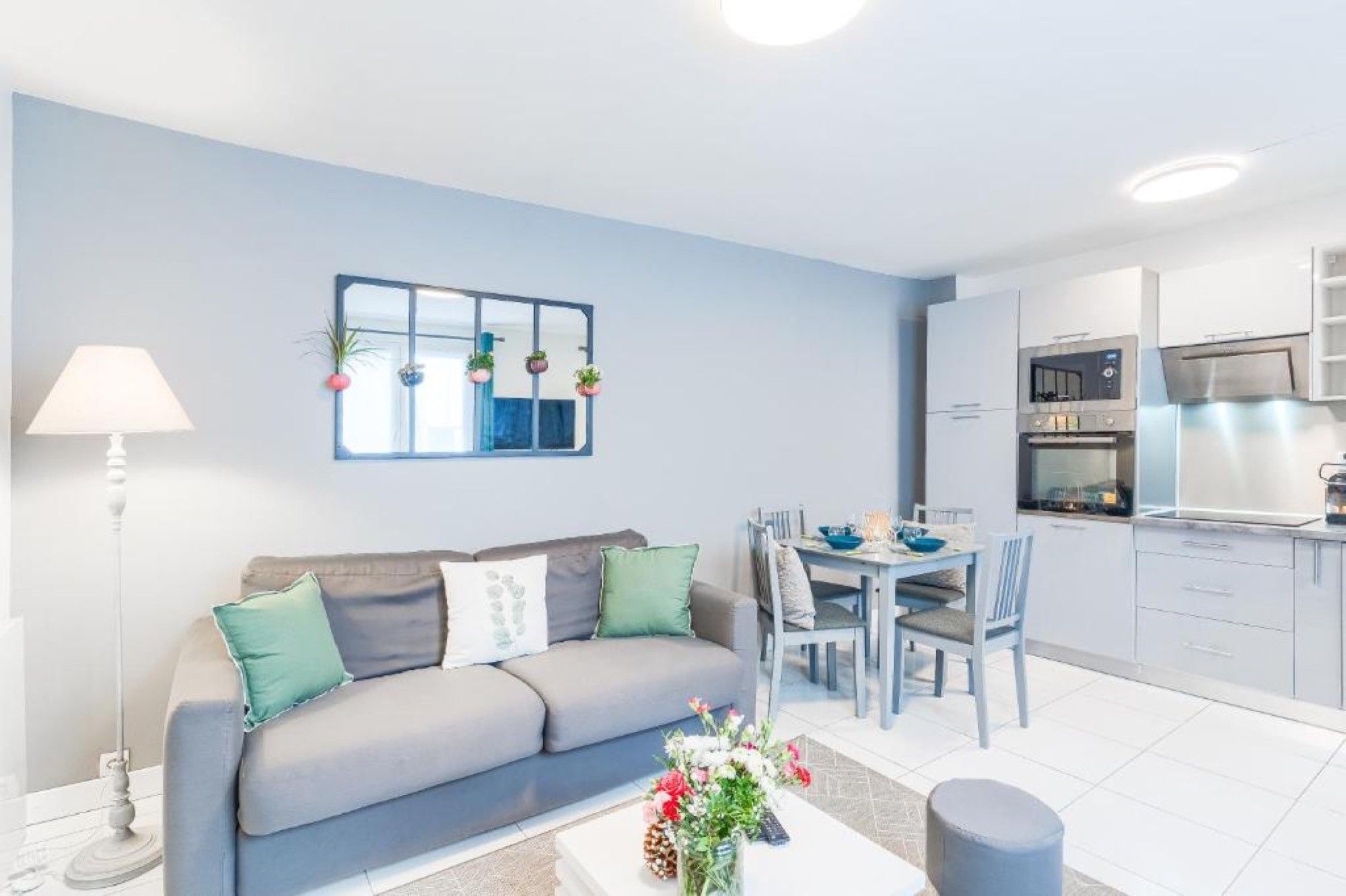 Un salon moderne comprenant un canapé, une table à manger avec des chaises, un coin cuisine avec des placards et des appareils électroménagers, et un miroir décoratif accroché au mur.