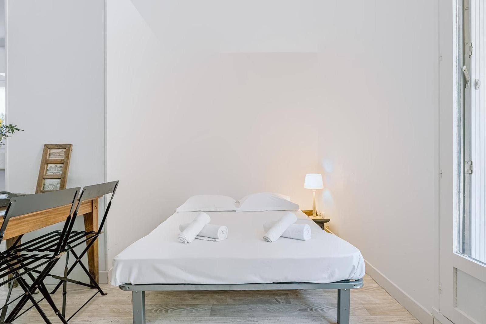 Een klein, netjes opgemaakt bed met witte lakens en kussens, een nachtkastje met een lamp, en een bureau met een stoel.