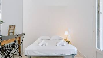 Een klein, netjes opgemaakt bed met witte lakens en kussens, een nachtkastje met een lamp, en een bureau met een stoel.