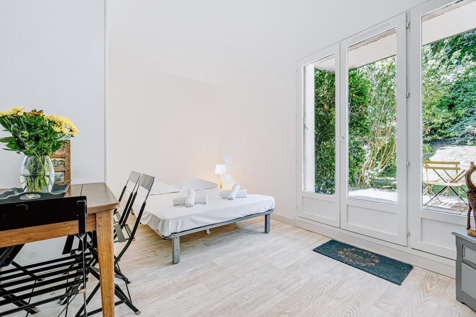 Een kamer met een bed, een tafel met stoelen, een vaas met bloemen en uitzicht op groen door een glazen deur.