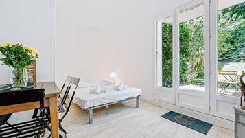 Een kamer met een bed, een tafel met stoelen, een vaas met bloemen en uitzicht op groen door een glazen deur.
