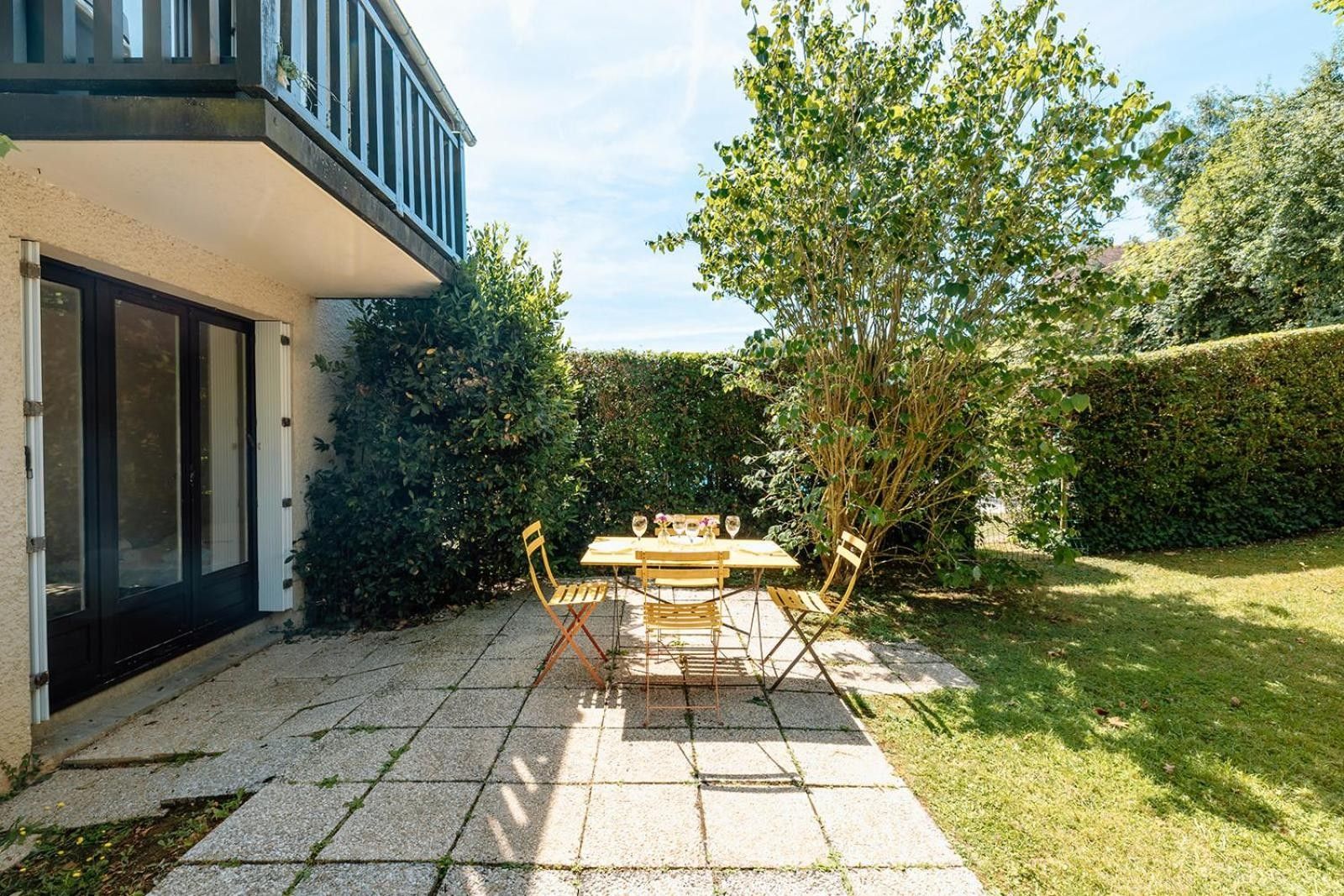 Een terras met een tafel en stoelen, een gebouw met balkon, en een keurig onderhouden haag.