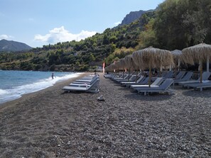 Ein Kiesstrand mit Liegestühlen und Reetdachschirmen, mit Blick auf eine ruhige See und eine gebirgige Kulisse.