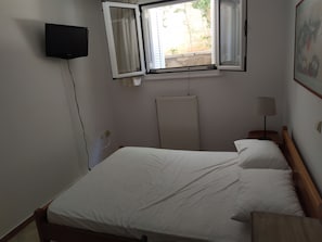 Ein Schlafzimmer mit einem Bett, einem Wand-Fernseher, einem Fenster mit Vorhängen, einem Heizkörper und einem Nachttisch mit Lampe.