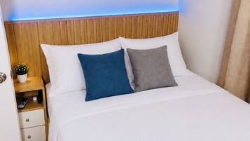 A neatly made bed with white bedding and two pillows, a wooden headboard, a bedside table with a plant, and a wall-mounted shelf with plants.