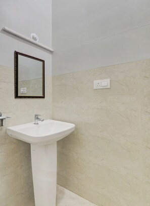 Un bany amb un lavabo pedestal blanc, un mirall fixat a la paret amb una estructura decorativa, i un interruptor de llum a la pared.
