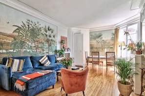 A living room with a blue sofa, a pink chair, and a dining area with a table and chairs. - Elegant Parisian Apartment minutes from Trocadero – Passy, 16th Arrondissement (Paris)
