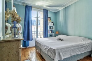 Une chambre à coucher avec un lit, une table de chevet, une fenêtre avec des rideaux, une bibliothèque et une lampe.