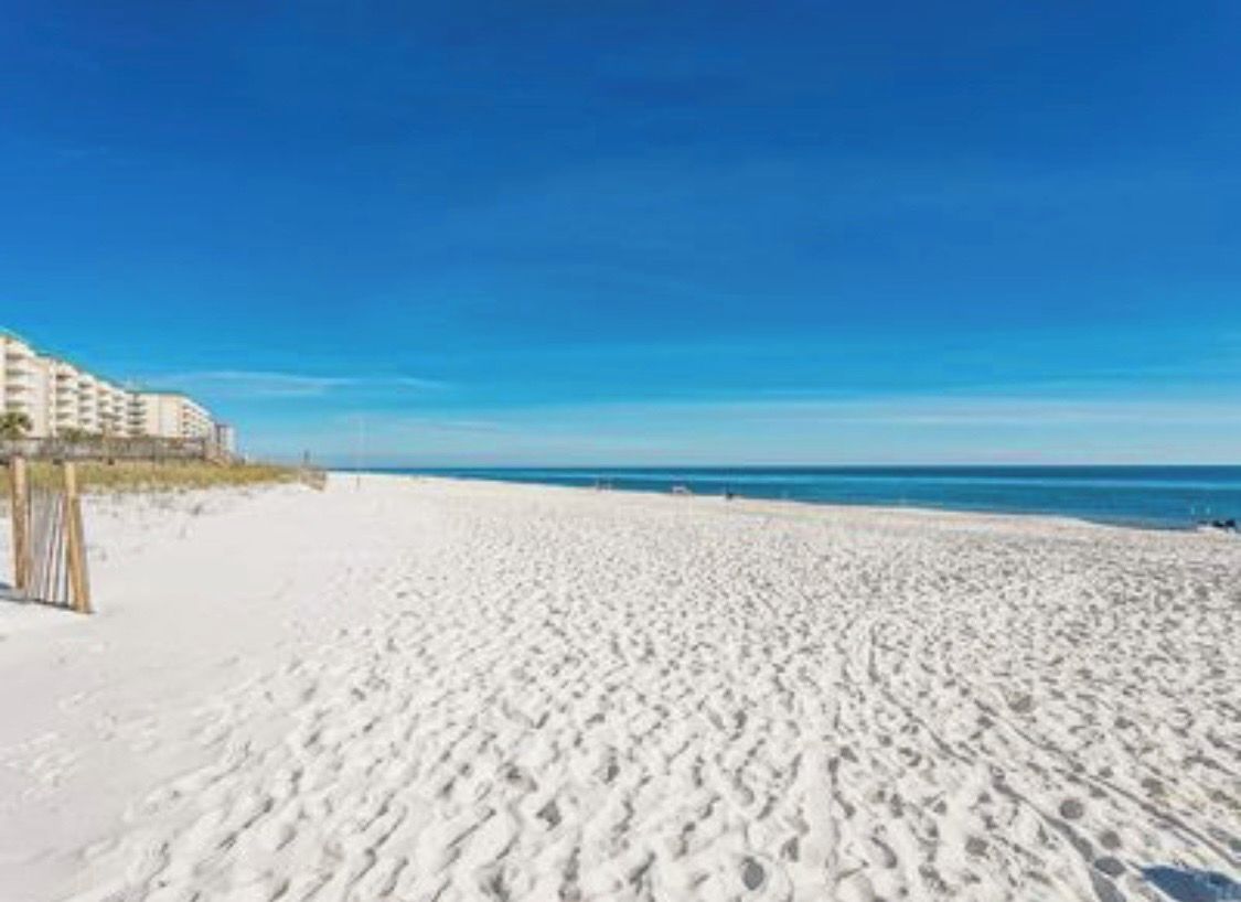 Une plage de sable avec des empreintes, un ciel dégagé et un bâtiment en arrière-plan.