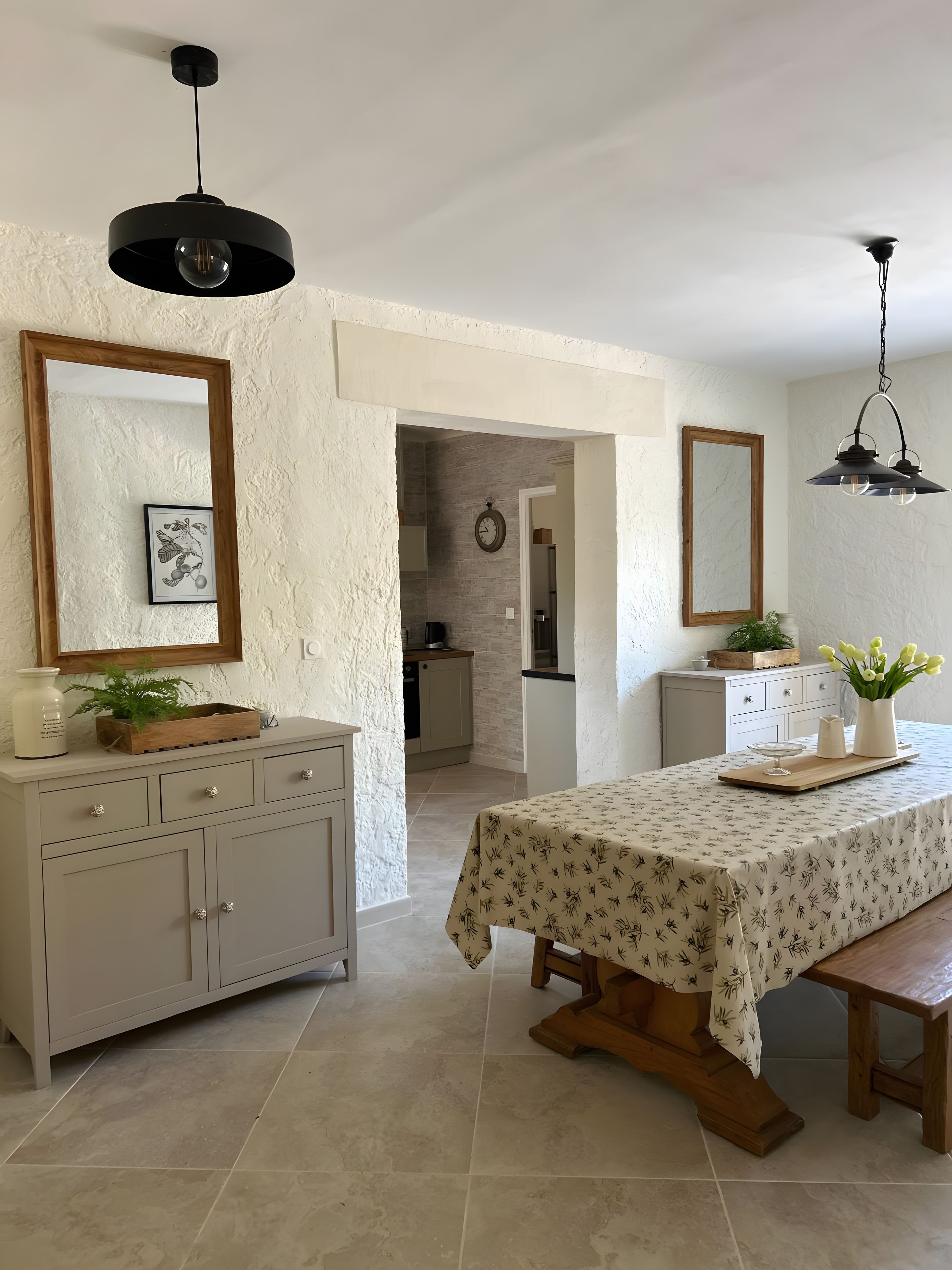 Un coin repas avec une table dressée pour deux personnes, un banc en bois et une cuisine en arrière-plan.