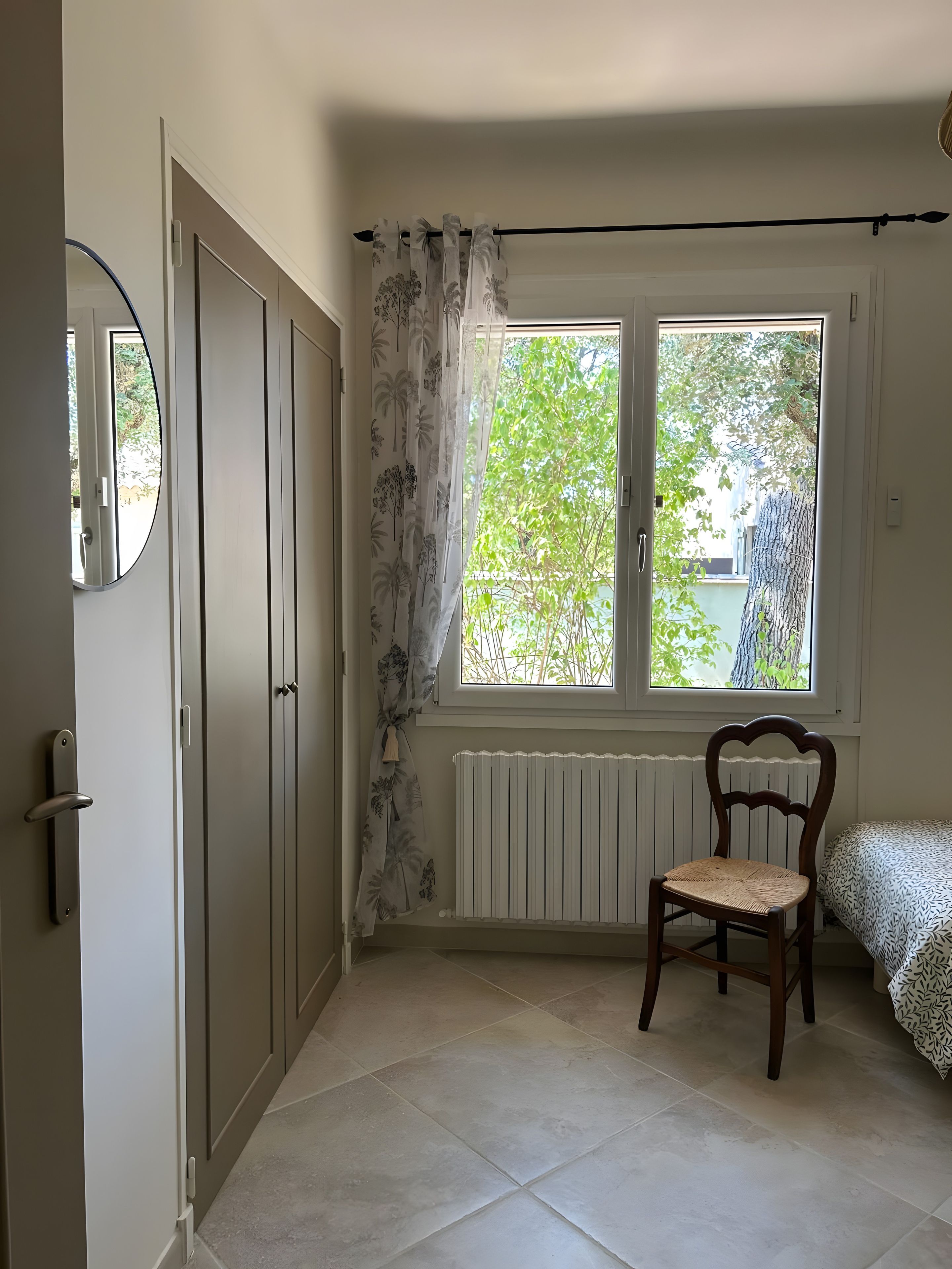 Une pièce avec une grande fenêtre, un radiateur, une chaise en bois et un lit avec une literie à motifs.