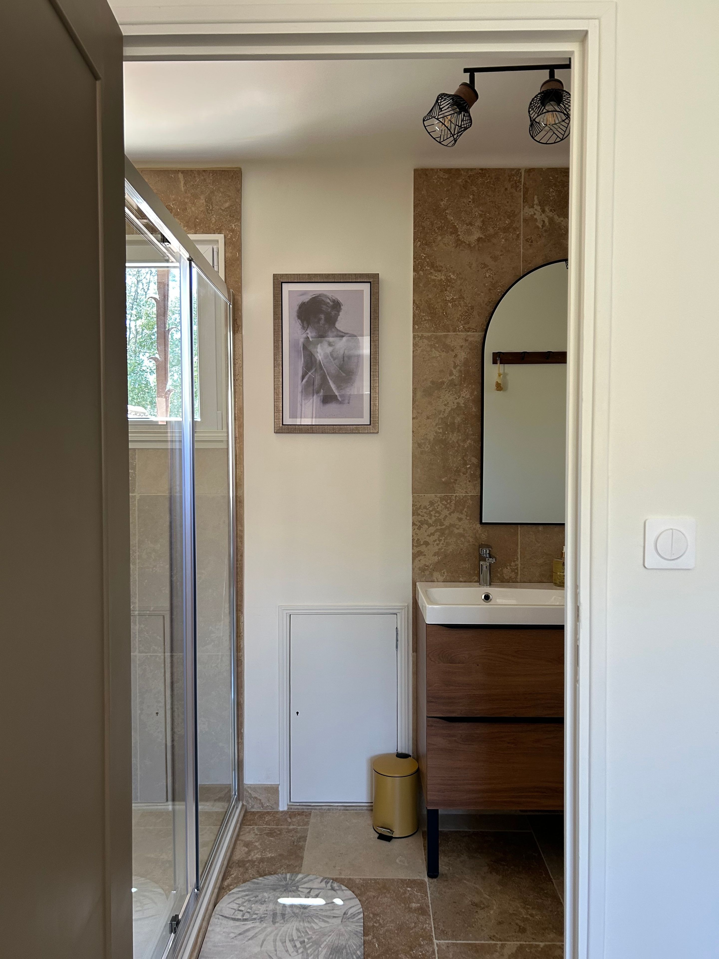 Une salle de bain avec une douche à parois de verre, un tableau encadré au mur, un lavabo avec un miroir et une poubelle.