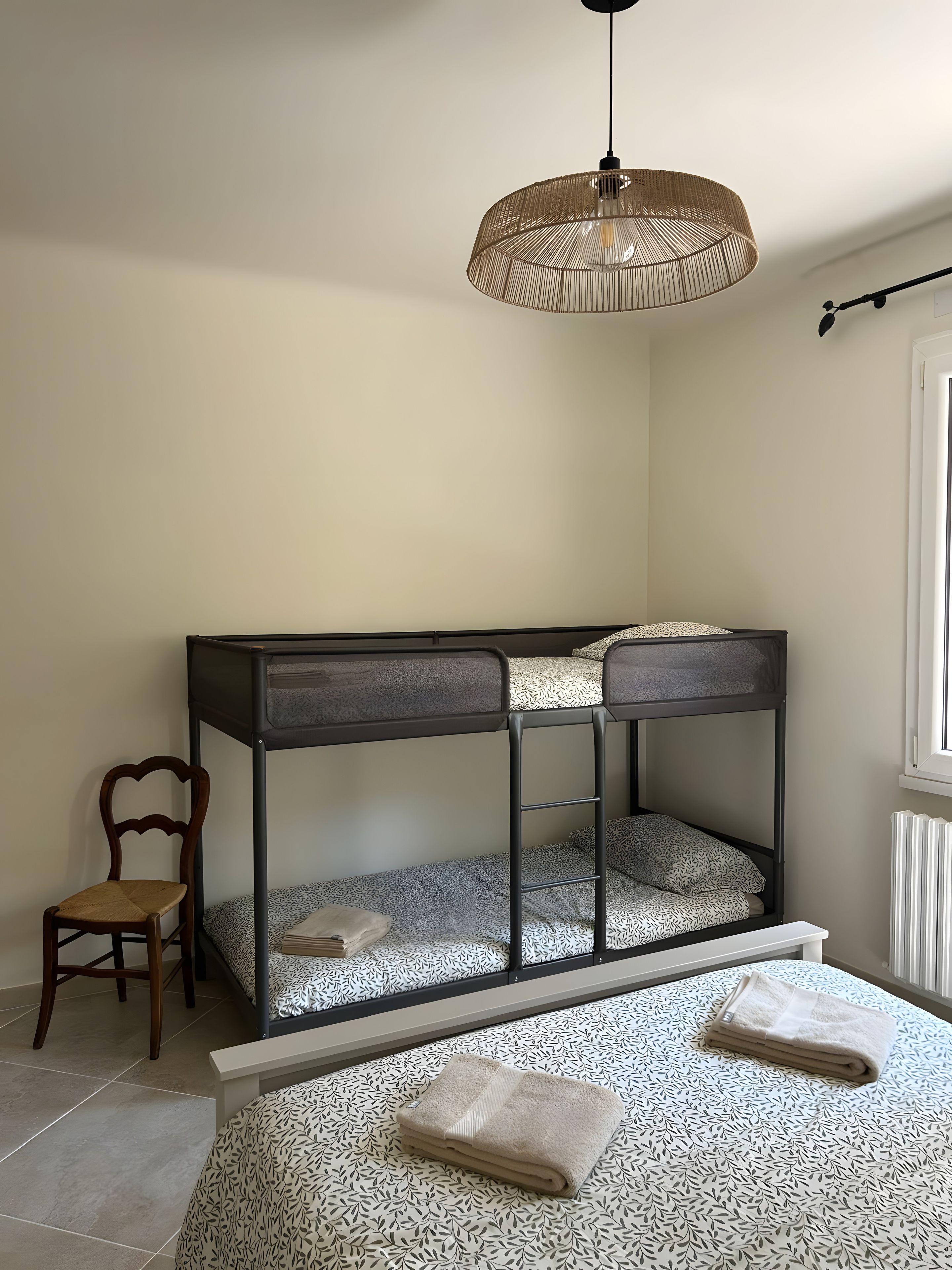 Un lit superposé avec une couvre-lit à motifs, une chaise en bois, un luminaire suspendu et un radiateur.
