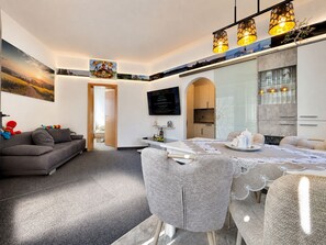 A modern living room with a sofa, dining table, and chairs, a kitchen in the background, and a TV mounted on the wall.