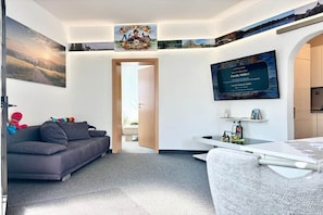 A modern living room with a grey sofa, a dining table, and a kitchen area visible through an open door. - Grossi's Haamit-Stüb'l (Schneeberg)