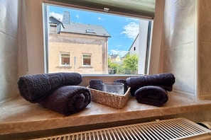 Ein Fenster mit Blick auf ein Gebäude, ein Fensterbrett mit aufgerollten Handtüchern und ein Korbwägelchen.