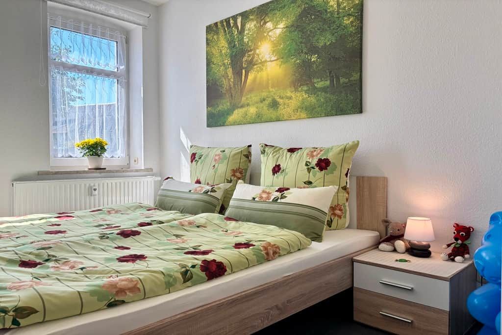 Ein Schlafzimmer mit einem Bett, einem Nachttisch, einem Fenster mit Vorhängen, einem Wandbild und einem kleinen Tisch mit einer Lampe und Teddybären.