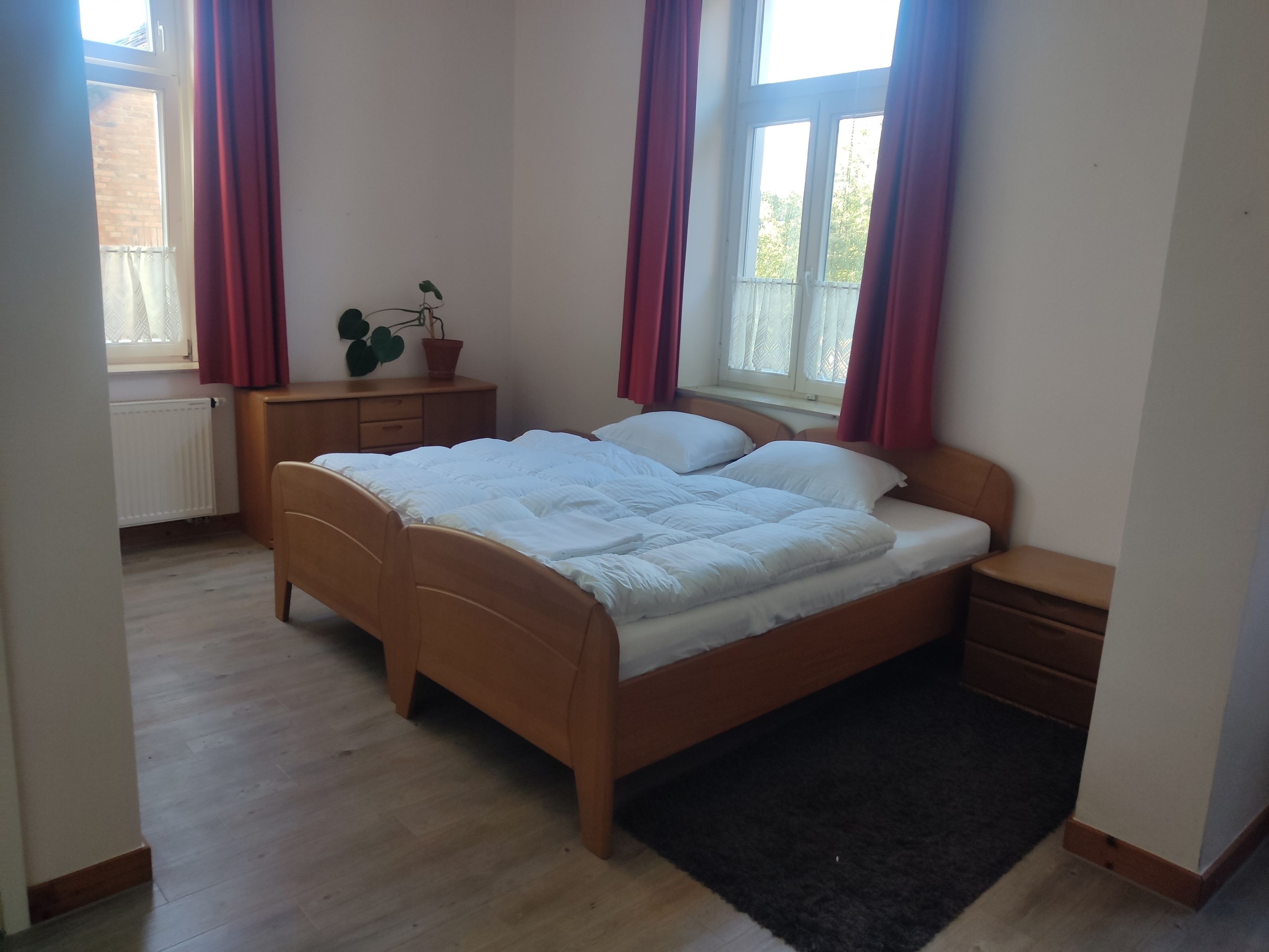 Un dormitorio con cama de madera, una mesita de noche, un radiador, una ventana con cortinas y una planta en maceta.