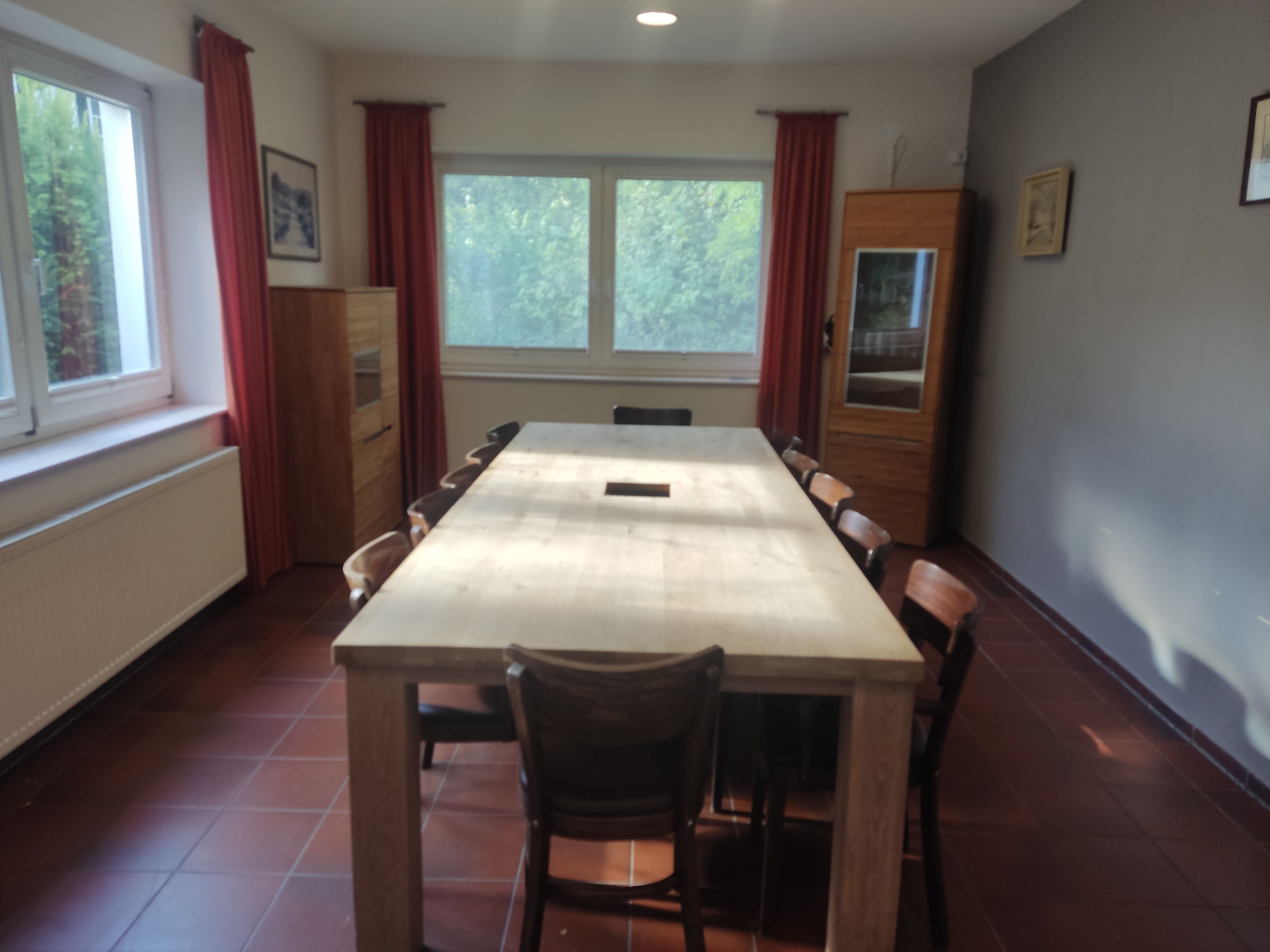 A dining room with a long table, chairs, a cabinet, and windows with curtains.